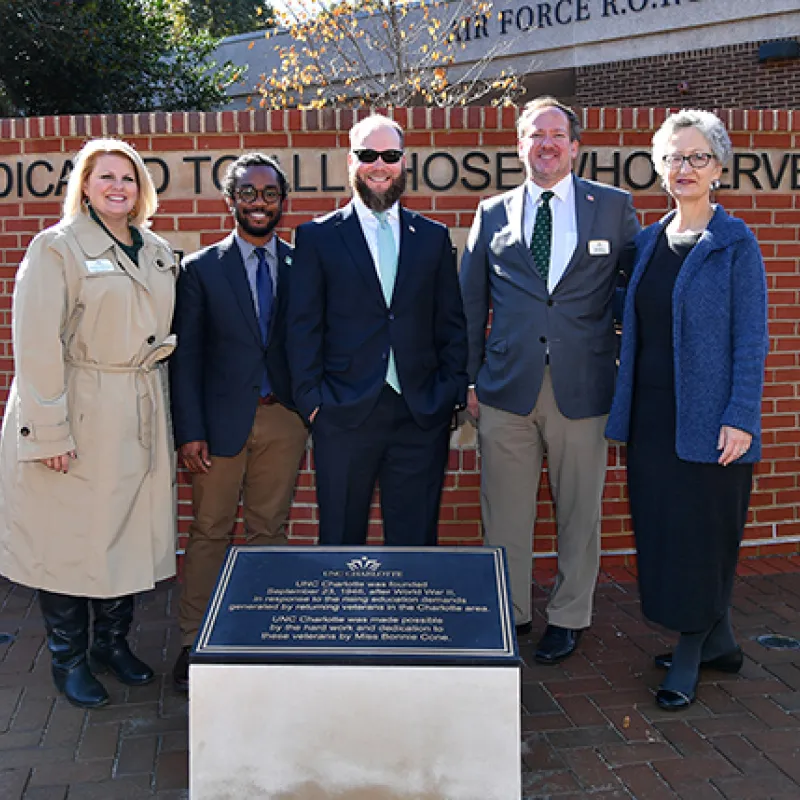 Group of five staff and students at dedication ceremony in front of brick memorial wall and plaque
